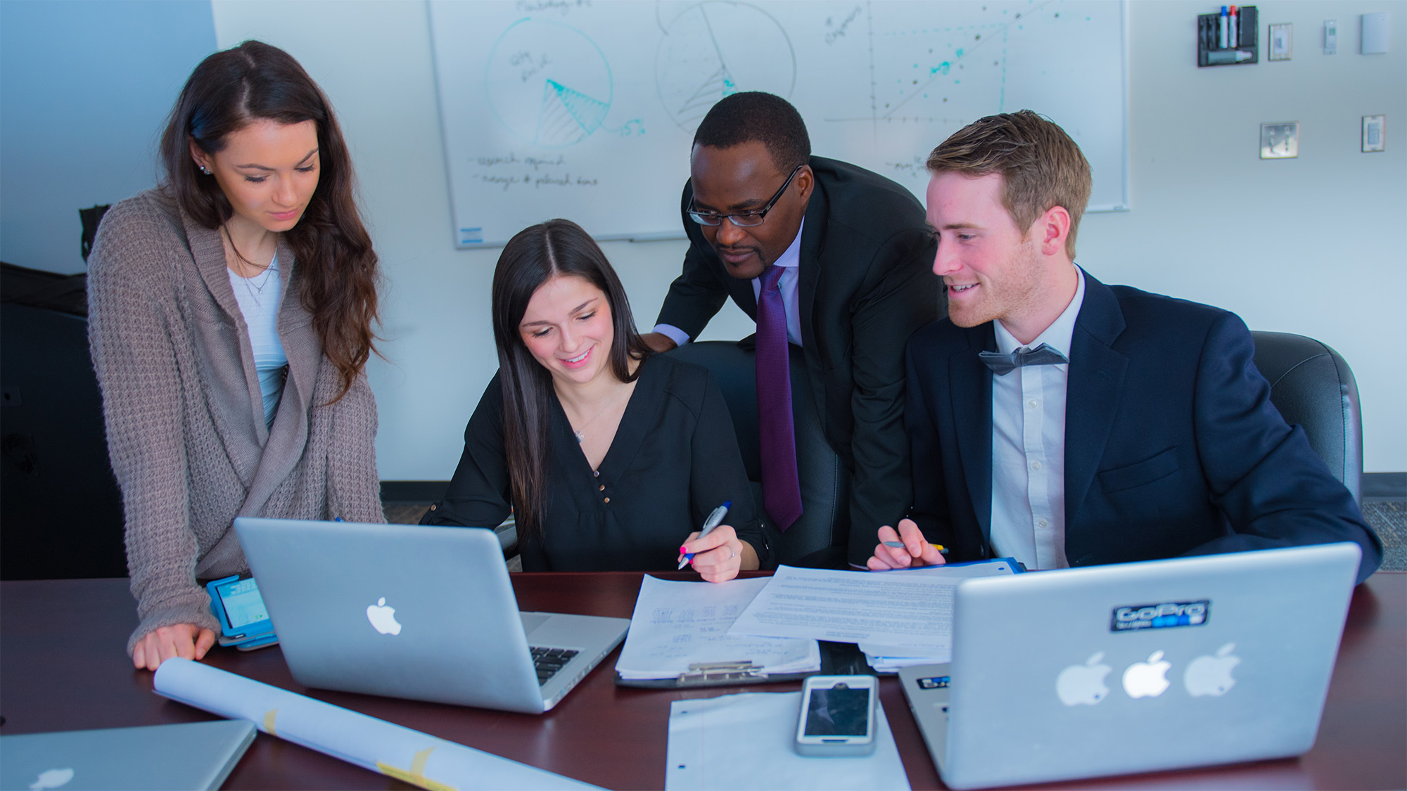 A group of students and a professor in business attire looking at a laptop in a work setting
