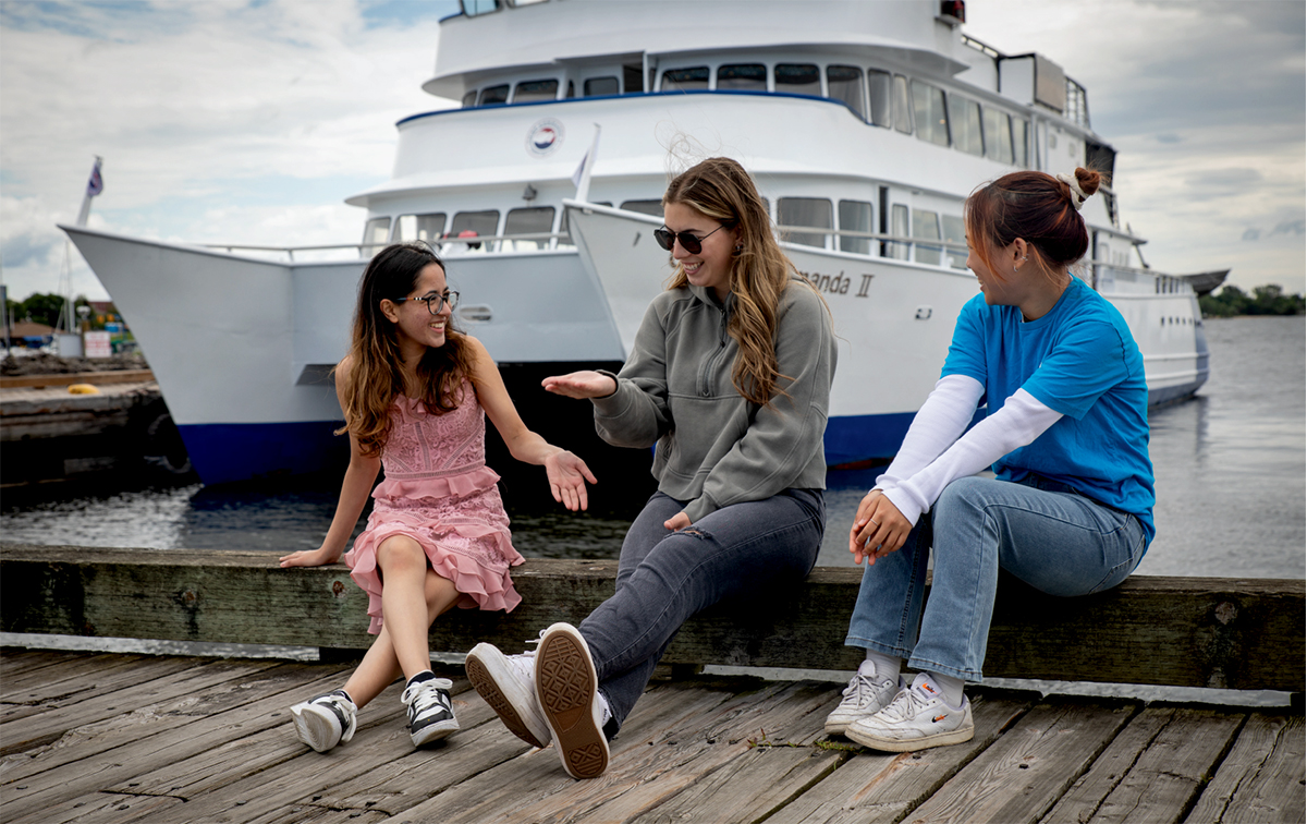 3 students sitting on the pier in front of the Chief Commanda II