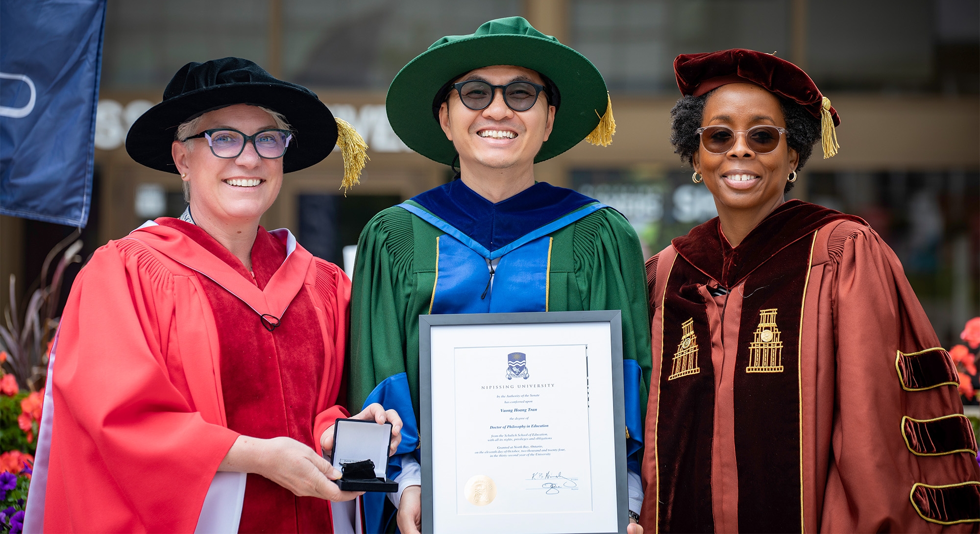 A graduate in green convocation gown holding their degree with 2 professors on both sides of him