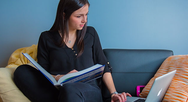 A student reading a book and going on her laptop on a sofa