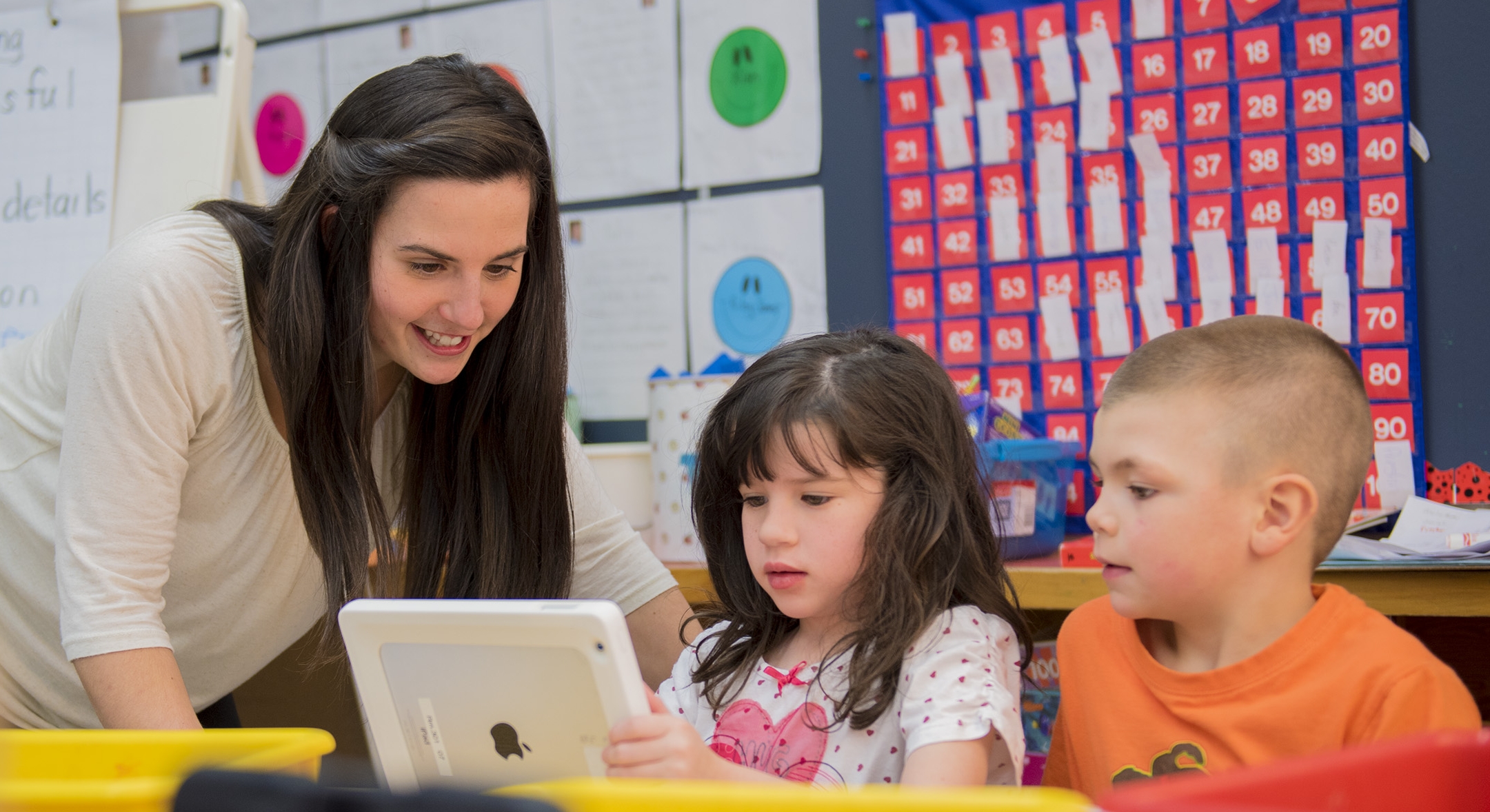 A student teacher helping 2 students with something on the ipad in a classroom setting