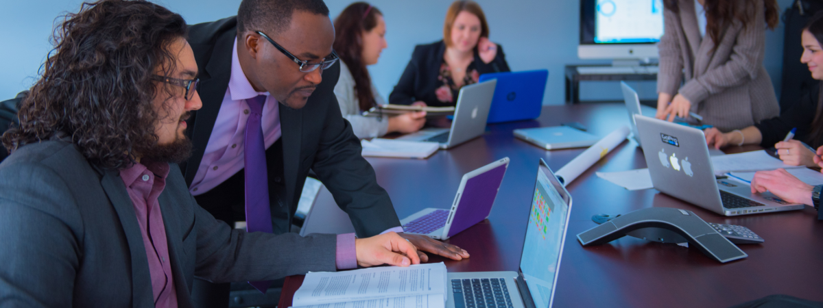 School of Business associates sit around a board table analyzing data on computers.