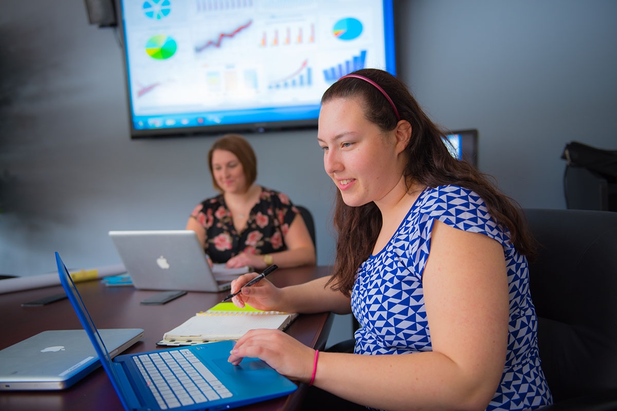 Two women work at macbooks with a monitor of charts on the wall above them.