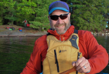 Terry Dubroy smiles at the camera from his canoe with a forested background.